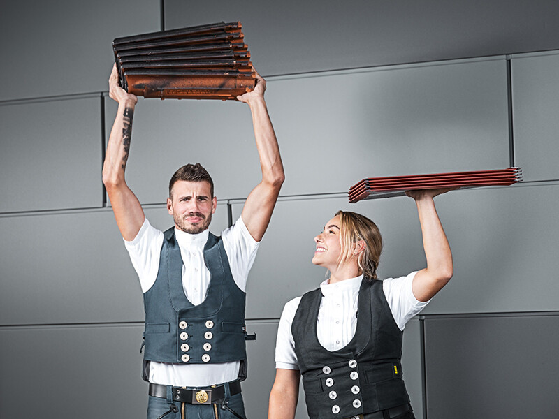 The image shows two people, each holding a stack of roofing material in their hands. The person on the left is holding a stack of roof tiles above their head, while the person on the right is holding a stack of aluminium roof panels, also above their head. Both are wearing traditional craftsmen's outfits with a waistcoat and a white shirt. The background is modern and minimalist, with grey walls. Both people appear focused and are smiling.
