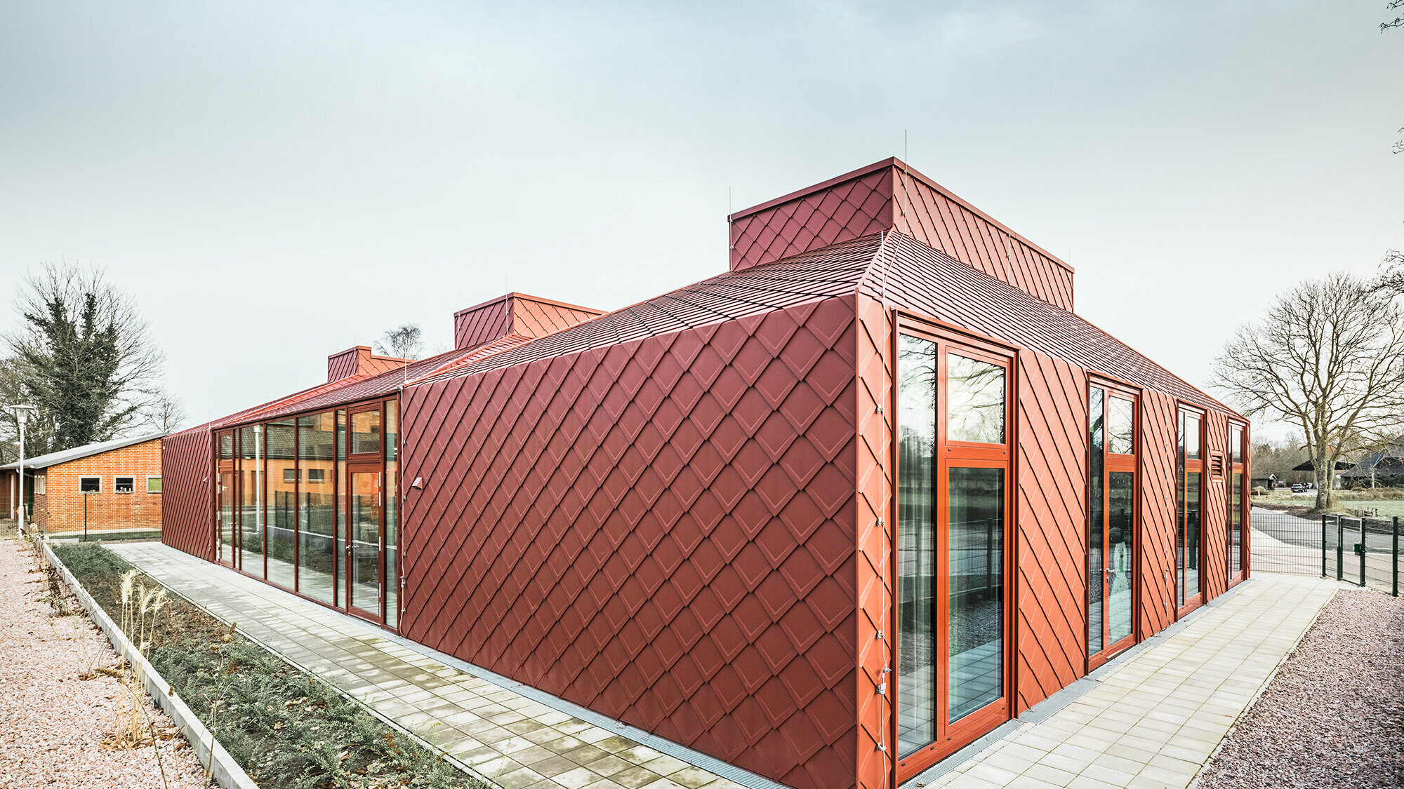Nowoczesny, parterowy budynek z czerwoną metalową fasadą i dachem firmy PREFA o rombowym wyglądzie, dużymi przeszkleniami sięgającymi od podłogi do sufitu oraz przejrzystą, nowoczesną architekturą, sfotografowany w świetle dziennym pod kątem od przodu.