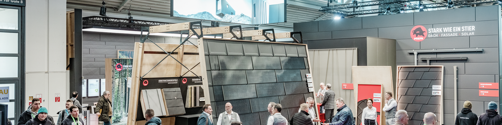 Der PREFA Messestand auf der BAU Messe, gekennzeichnet durch den Slogan „Stark wie ein Stier – Dach, Fassade, Solar“ und eine große hängende Werbefläche. Im Zentrum stehen präsentierte Aluminium-Dach- und Fassadensysteme sowie innovative Montagelösungen. Besucher tauschen sich mit PREFA Mitarbeitern an roten Beratungstischen aus, während weitere Exponate und Infotafeln im Hintergrund zu sehen sind.