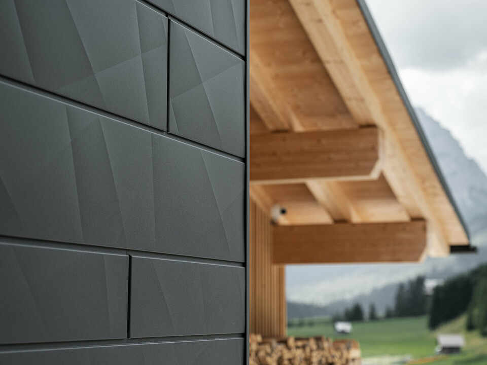 Close-up of the Siding.X façade panels in P.10 black from PREFA on Casa Hoffe in Sappada, Italy. The aluminium façade system has been skilfully installed in a modern, abstract design that forms an attractive contrast to the visible wooden elements of the roof structure. The innovative façade solution blends seamlessly into the alpine ambience and represents the combination of functionality, durability and aesthetic building culture.