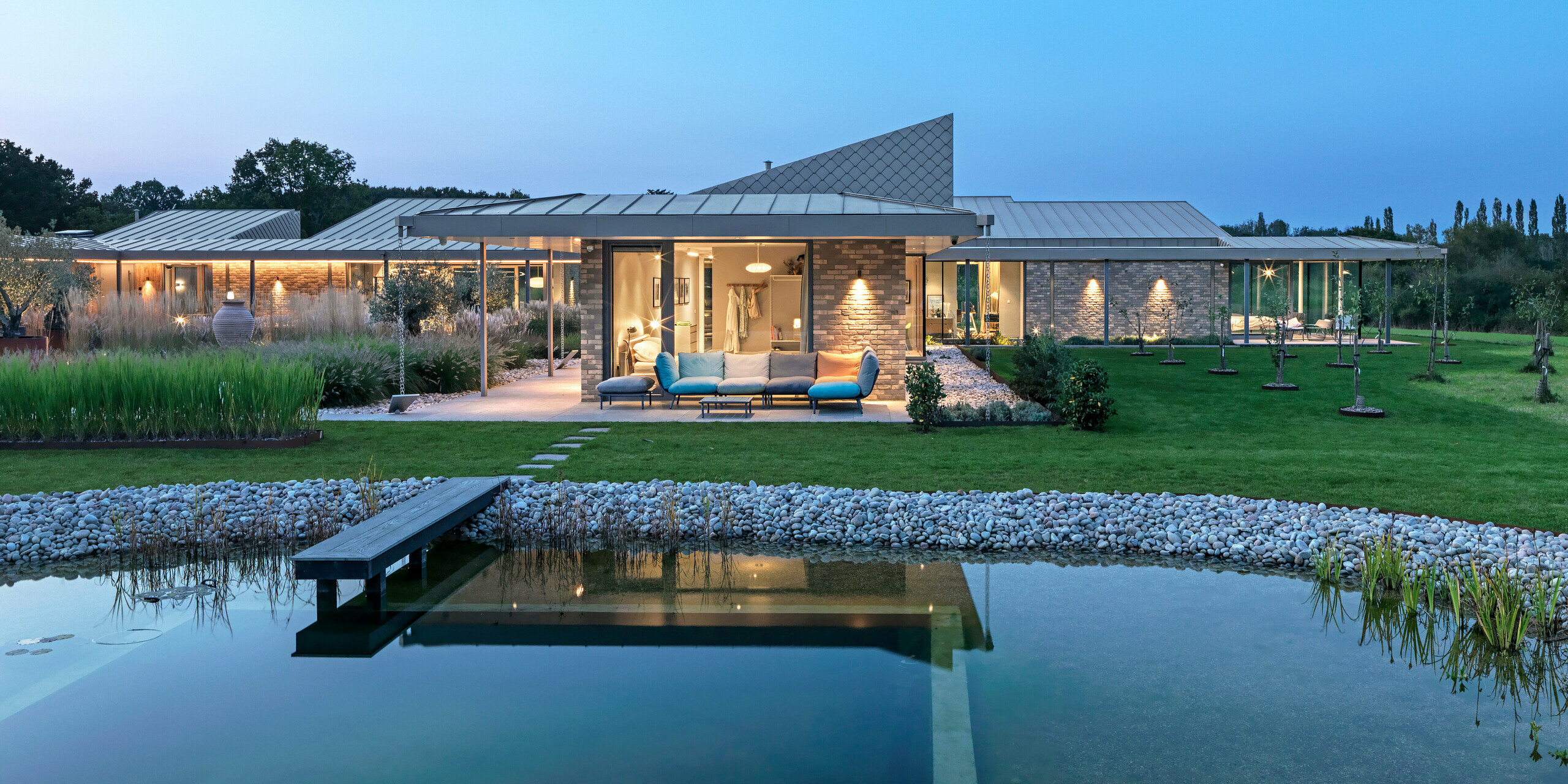 Evening view of a dream home in West Sussex with standing seam roof and façade accents made from PREFA rhomboid façade tiles 20 × 20 in P.10 bronze. In the foreground is a natural swimming pool with a wooden deck, behind which is a spacious terrace with lounge furniture and an illuminated veranda. The clean lines of the architecture and the high-quality PREFA products create an elegant combination of design, functionality and sustainability.