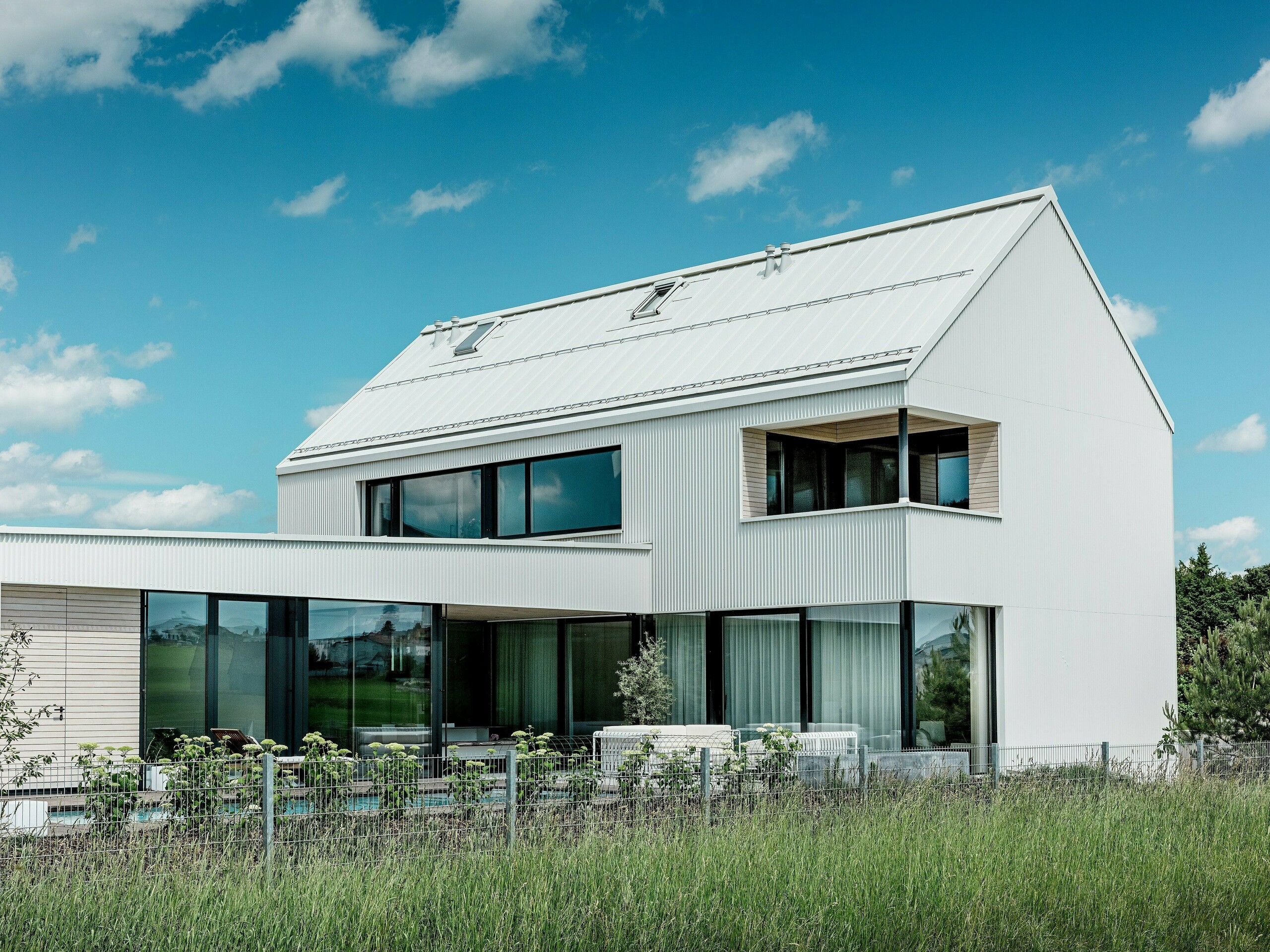 Dieses beeindruckende Einfamilienhaus in Freistadt besticht durch seine elegante Architektur, bei der reinweißes PREFALZ-Aluminium für Fassade und Dach verwendet wurde. Die zickzack gefaltete Fassade schafft ein modernes, geometrisches Erscheinungsbild, das sich harmonisch in die natürliche Umgebung einfügt. Große Fensterflächen bringen nicht nur viel Tageslicht in den Innenraum, sondern bieten auch einen weiten Blick auf die umliegende Landschaft. Das steile Satteldach und der moderne Balkon mit Holzverkleidung unterstreichen das minimalistische Design und die nachhaltige Bauweise des Hauses. PREFALZ sorgt dabei für optische Beständigkeit und Witterungsresistenz und garantiert eine lange Lebensdauer bei geringem Pflegeaufwand.