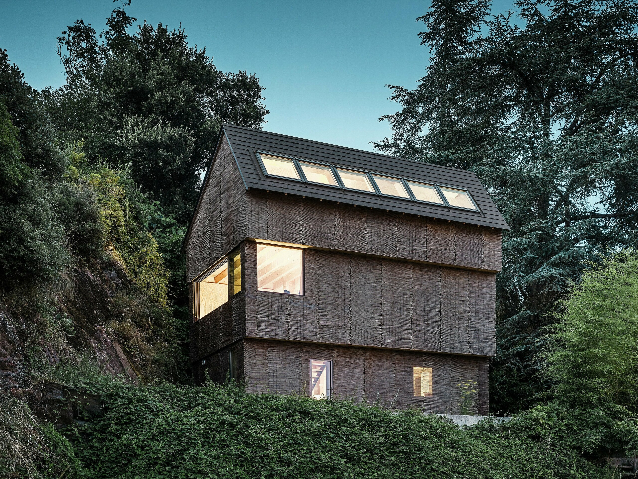 Nachtansicht eines modernen Hauses im Westen Frankreichs mit PREFA-Dachschindeln in der Farbe P.10 Walnussbraun, geräumigen Dachfenstern und einer Fassade aus nachhaltigem Weidenholz.