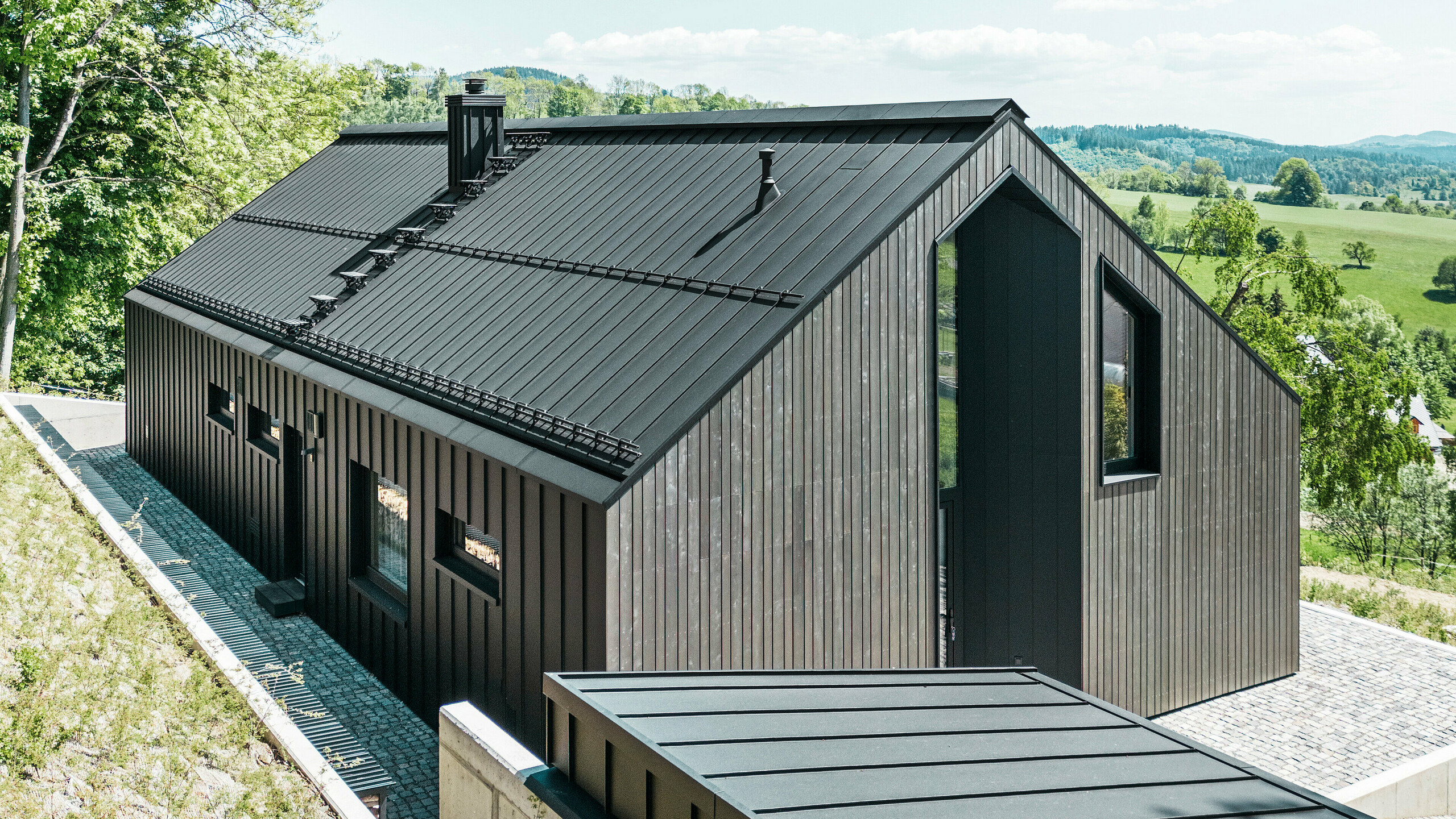 Dunkles Holzmassivhaus mit PREFALZ in P. 10 am Dach und der Rückseite des Gebäudes. Vor dem Haus steht ein kleines Nebengebäude mit einer Aluminiumhülle aus PREFALZ in P.10 Schwarz. Im Hintergrund ein strahlend blauer Himmel und Wald.