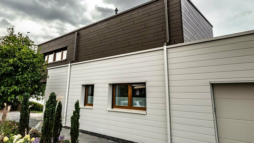 Das Bild zeigt ein Haus, das mit Prefa Siding in zwei Farben verkleidet wurde: der obere Teil der Fassade ist in einem eleganten Braunton gehalten, während der untere Bereich in Prefaweiß ausgeführt ist. Passend dazu verlaufen auch die Ablaufrohre in denselben Farben. Diese Kombination verleiht dem Gebäude ein modernes und stilvolles Aussehen.