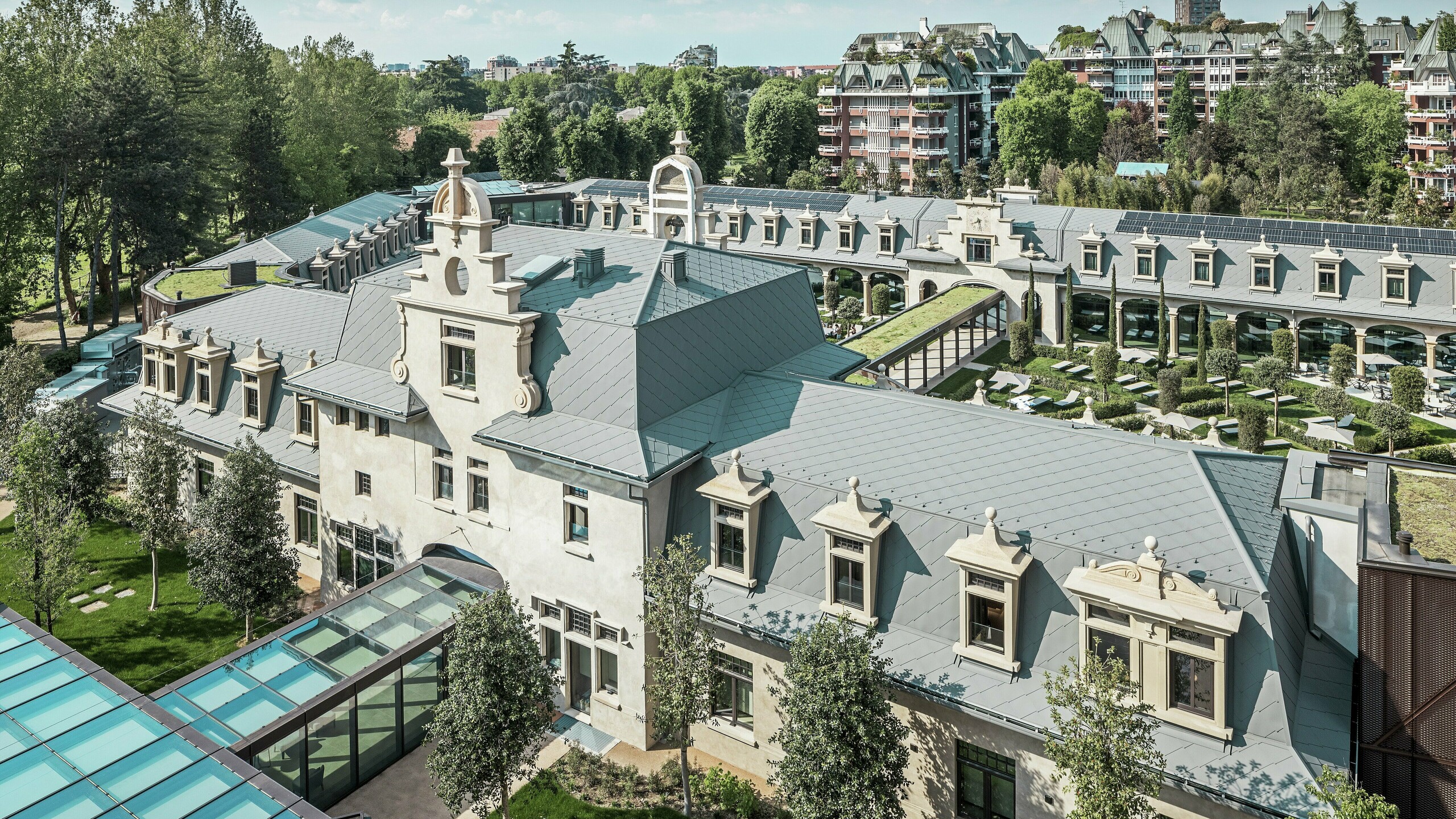 Overall view of the impressive ‘De Montel – Terme Milano’ complex with its listed Art Nouveau architecture and modern wellness ambience. The large-scale roof covering with PREFA rhomboid roof tile 44 × 44 in P.10 light grey gives the complex an elegant uniformity. Integrated PV modules, green roofs and glass connections between the buildings reflect the demand for sustainability and contemporary design in an urban context.