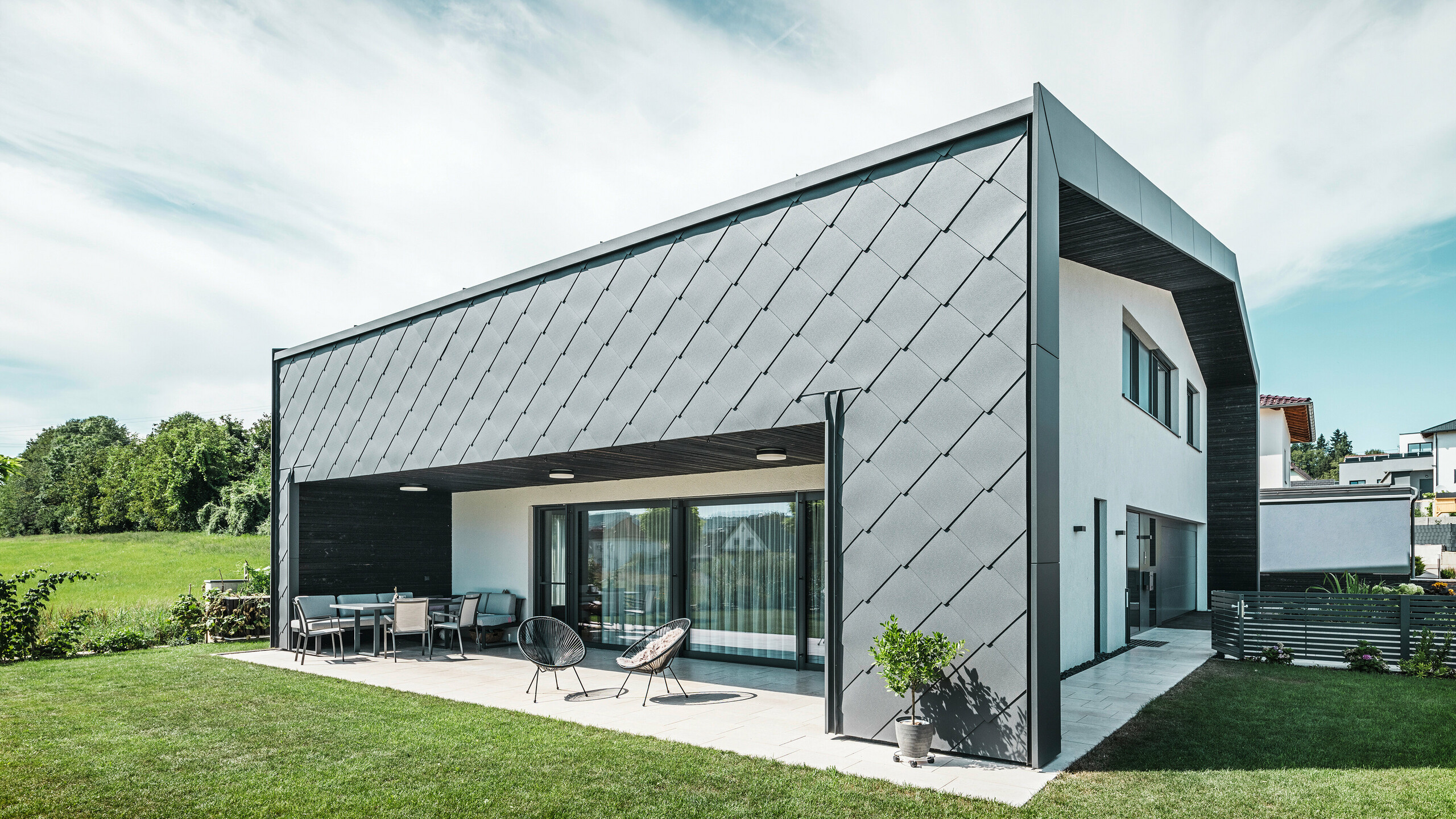 Well-kept garden area of a detached house in Aschach. The building is stylishly clad with dark grey PREFA aluminium rhomboid façade tiles 44 × 44. The covered terrace offers a cozy place to sit with a view of the lush green lawn. The elegant aluminium cladding in combination with white plaster surfaces ensures a modern and weather-resistant architecture. Large windows allow plenty of daylight into the interior, creating an open, light-filled atmosphere.