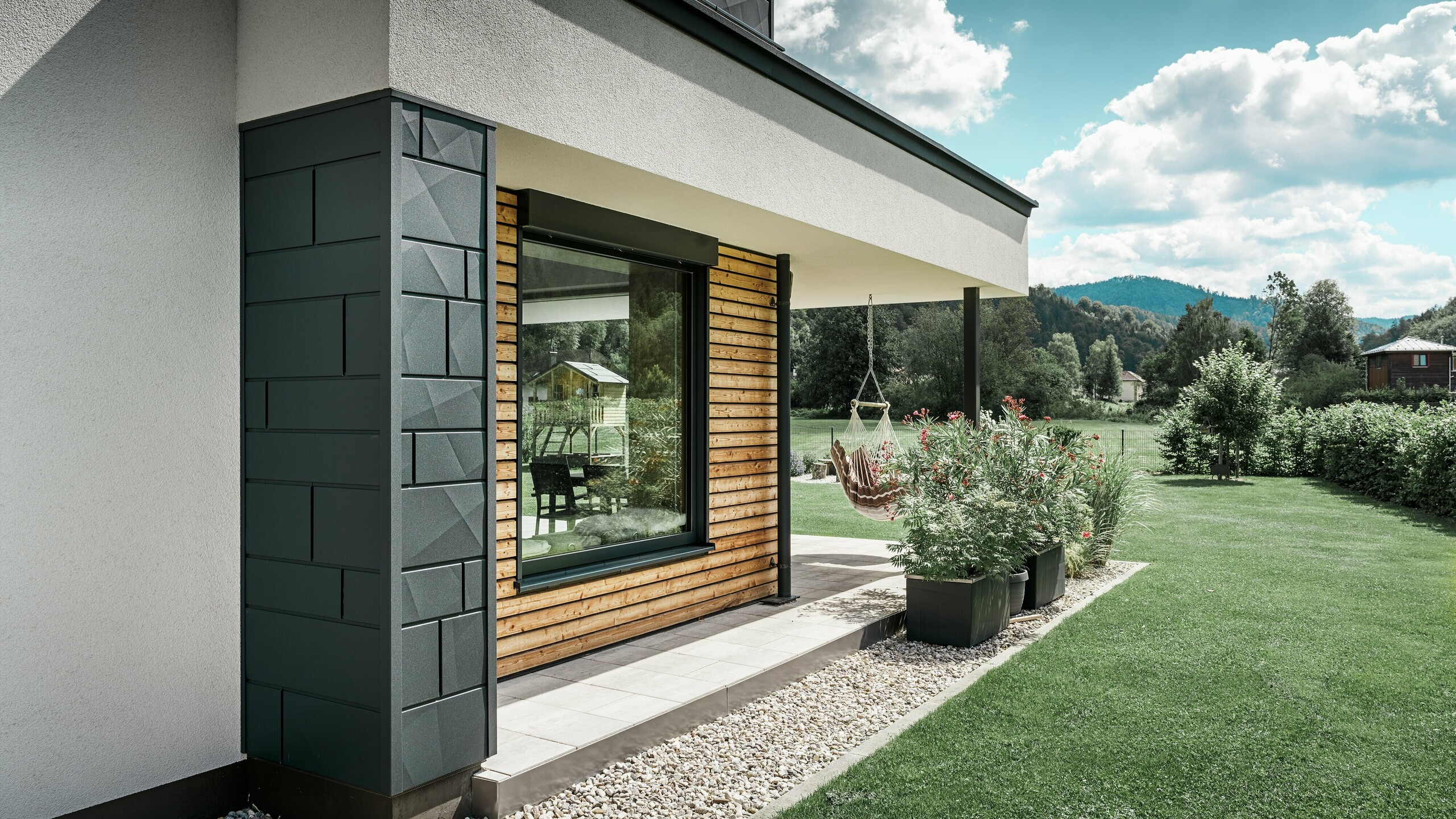 Partial view of a newly built detached house. Here, different materials were used on the façade. The white plaster façade is highlighted with PREFA Aluminium Siding.X in anthracite and horizontal wooden panels that surround a large window. There is a hanging chair on the terrace.