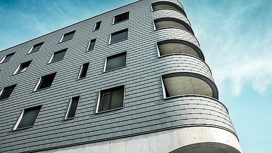 Detailaufnahme des Bernerhauses in Buchs mit markant gerundeter Gebäudeecke und fließenden Balkonöffnungen. Die Fassade aus PREFA Wandschindeln in P.10 Hellgrau legt sich wie eine präzise verarbeitete Aluminiumhaut über die klar strukturierte Kubatur. Die schuppenartige Oberfläche betont die weichen Radien und sorgt für ein lebendiges Licht- und Schattenspiel. Dank des leichten, witterungsbeständigen und langlebigen PREFA Aluminiums entsteht eine homogene, wartungsarme Gebäudehülle – technisch anspruchsvoll und architektonisch ein echter Eyecatcher.