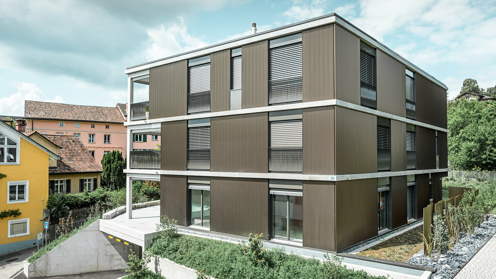 The striking aluminium façade of this apartment building in Schaffhausen features the elegant PREFA serrated profile in the colour Colinal. The vertical arrangement of the extruded aluminium profiles makes the building appear more compact and gives the façade structure a harmonious balance. The sophisticated design blends tastefully into the surroundings and gives the building a modern elegance that is both visually appealing and functional. The perspective shows the apartment building from the front with the garage entrance. Trees, other buildings and a church tower can be seen in the background.