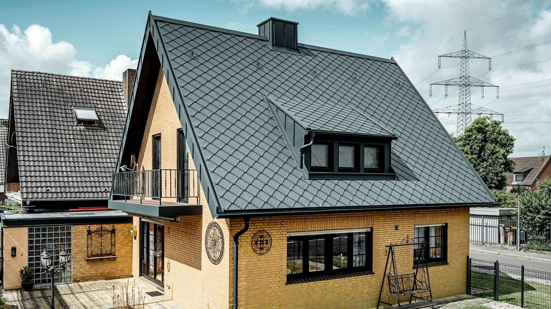Detached house made of yellow clinker bricks after roof renovation with PREFA rhomboid roof tile 29 × 29 in dark grey; newly covered, steep gable roof with diamond pattern, integrated dormer and chimney, terrace and garden in the foreground.