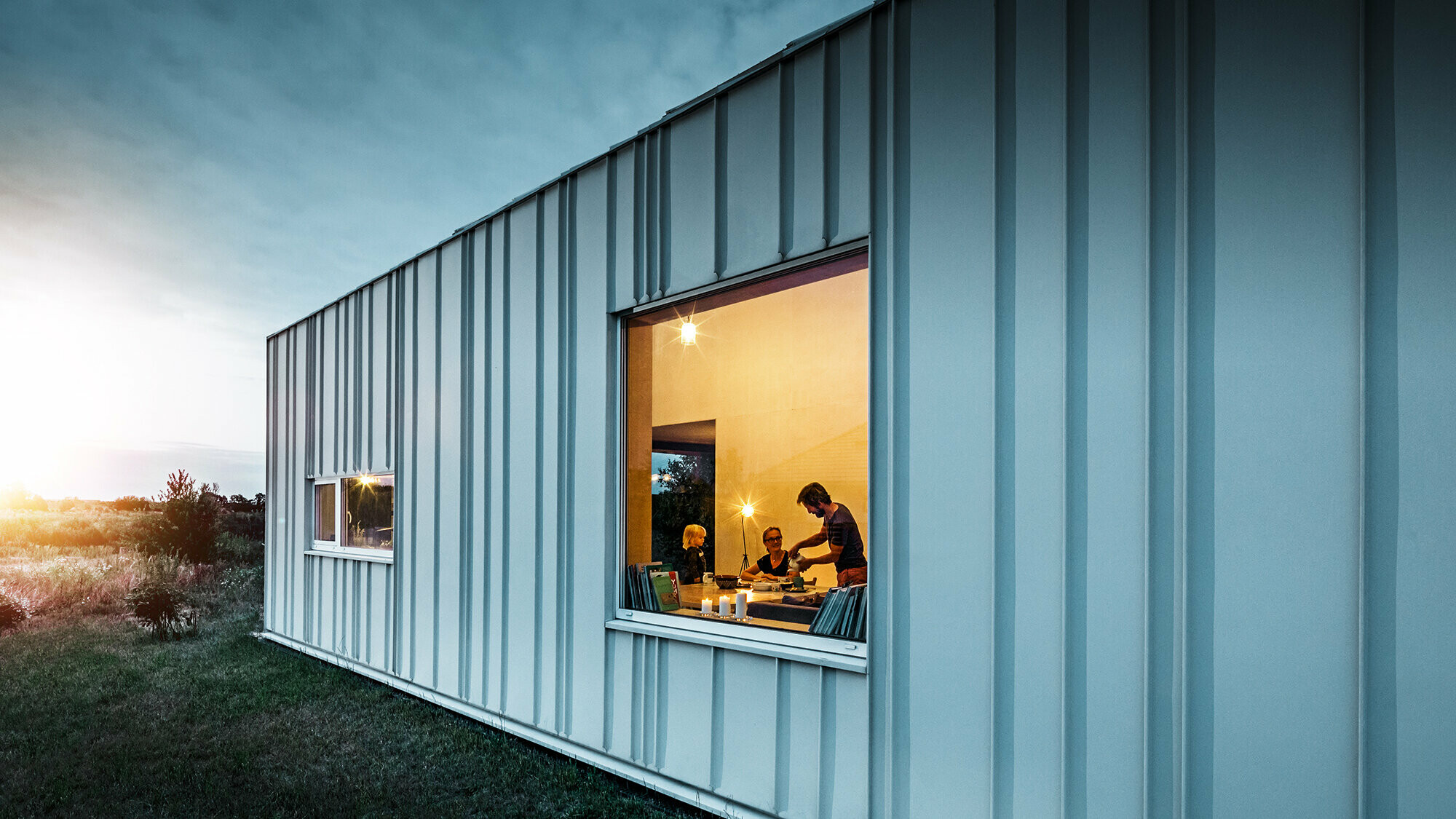 Angled standing seam façade with PREFALZ in different panel widths in P.10 Prefa white photographed at dusk. Across the dining room, you can make out a family having dinner. 