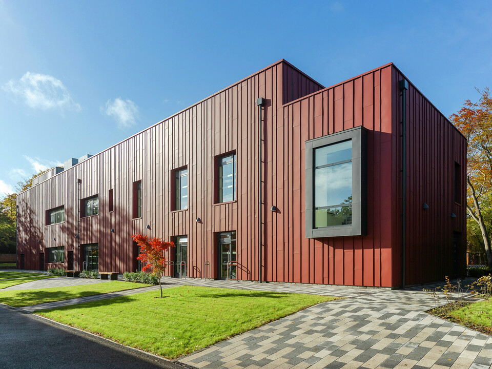 Modern architecture with a robust aluminium façade: the Soldiers' Centre in Pirbright presents an elegant standing seam cladding made of PREFALZ in P.10 oxide red, which blends harmoniously into the green surroundings. The prominent, cantilevered window frames in P.10 anthracite set architectural accents. The paved path and the well-kept green space emphasise the inviting design of the building. Sustainable and aesthetic construction with PREFA aluminium products.