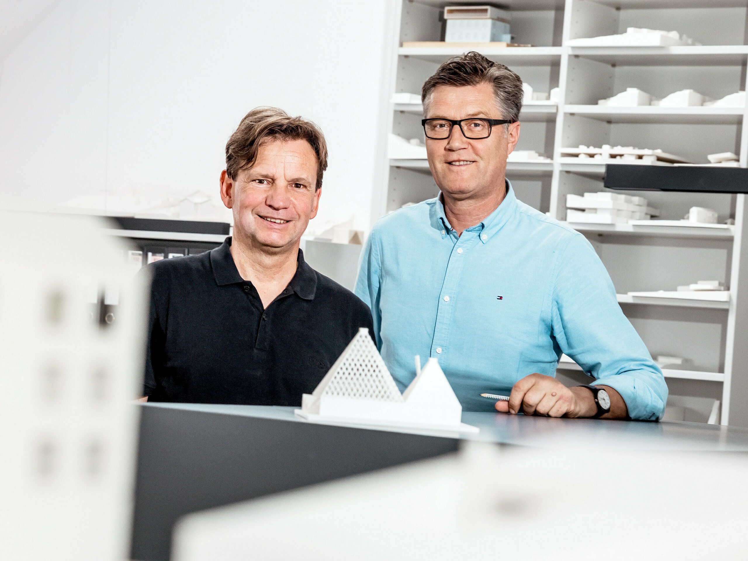 Auf dem Bild sind die Architekten von Schneider Lengauer Pühringer zu sehen, wie sie stolz in ihrem Büro vor einer Sammlung von Architekturmodellen posieren. Sie haben das moderne Einfamilienhaus in Freistadt mit dem Projektnamen „One Shade Of White” entworfen und sind für ihre innovative und ästhetische Herangehensweise an Architektur bekannt. Die Modelle im Hintergrund spiegeln ihre kreativen Konzepte wider und zeigen ihre Fähigkeit, moderne Designideen mit funktionalen Lösungen zu verbinden. Beide tragen legere Kleidung, was die entspannte, aber gleichzeitig professionelle Atmosphäre in ihrem Arbeitsumfeld unterstreicht.