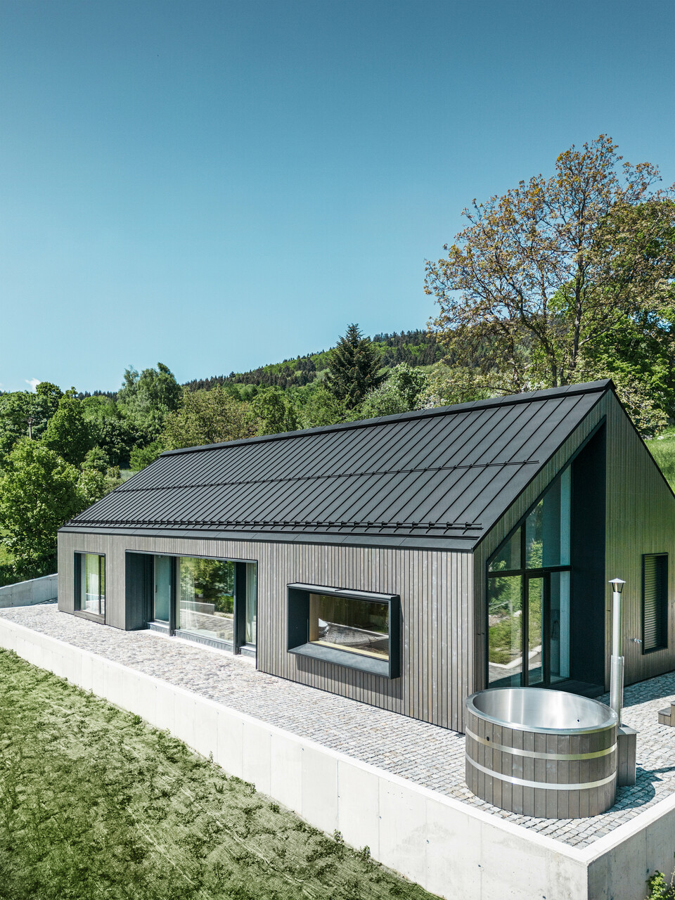 A modern wooden house in Kuncice (CZ) was equipped with large windows. The dark wooden house has a PREFALZ (standing seam) roof system in the colour P.10 black. In the rear outdoor area, there is a hot tub with wooden panelling.