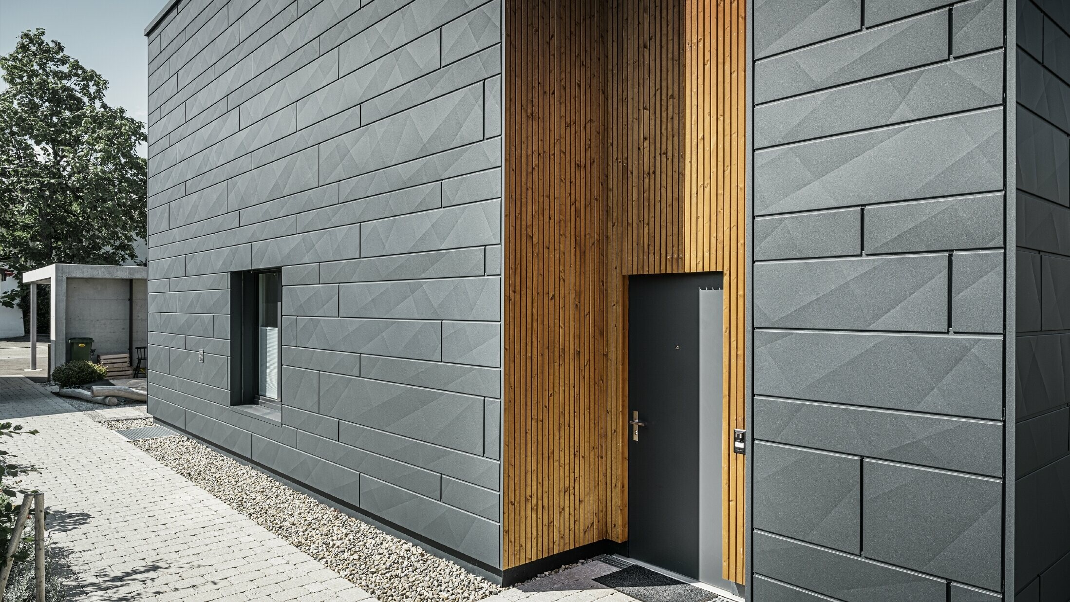 Zu sehen ist der Eingangbereich des modernen Einfamilienhauses. Die Fassade ist mit PREFA Siding.X verkleidet, der Türbereich ist aus Holz.