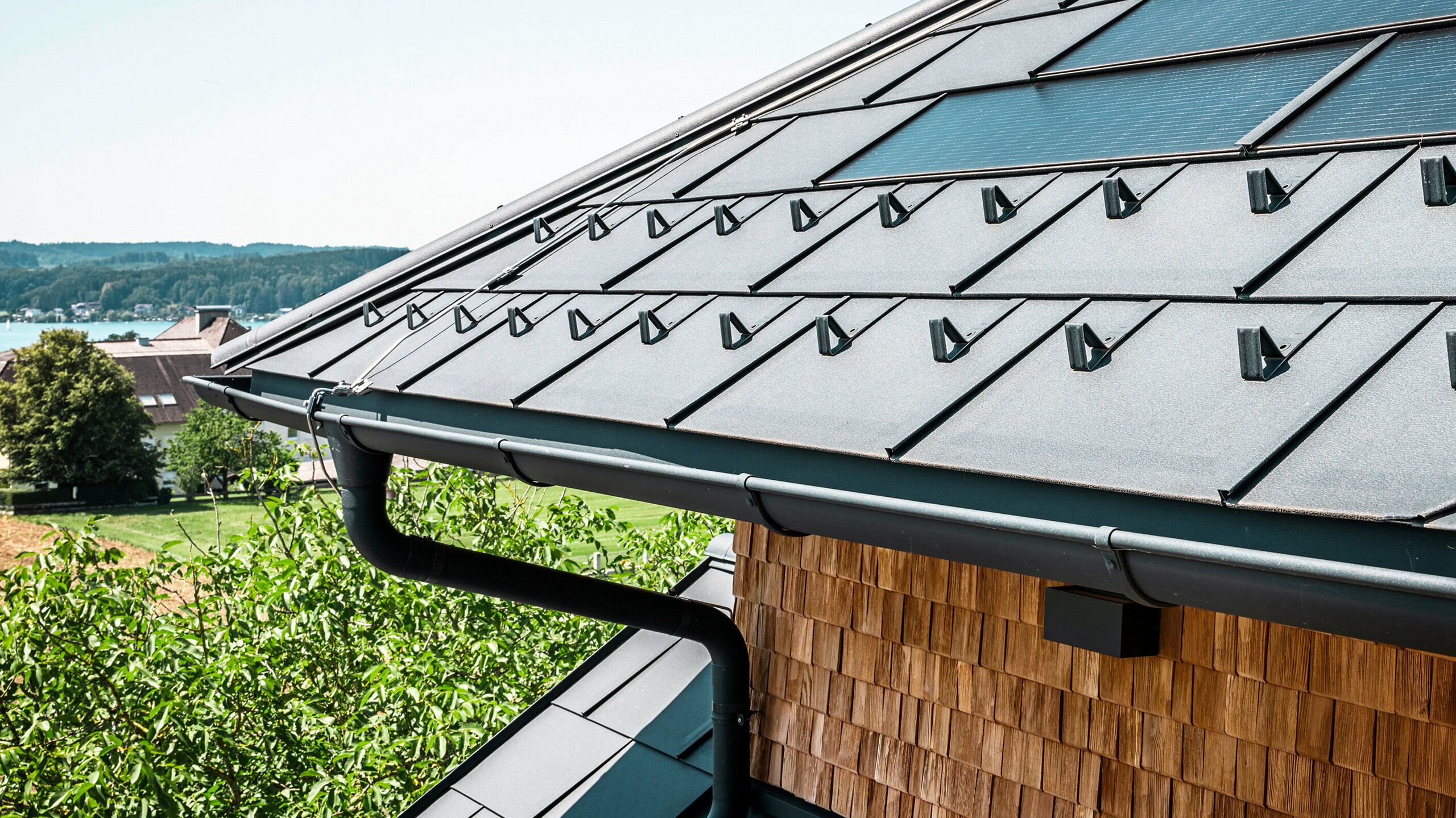The Zenz'n Stub'n in Schörfling am Attersee presents a newly renovated roof with innovative PREFA solar roof tiles and durable PREFA R.16 roof tiles in P.10 anthracite. The stylish aluminium roof combines modern design with sustainable energy generation. The roof drainage system, consisting of downpipes and half-round gutters in P.10 anthracite, blends harmoniously into the overall picture and emphasises the high-quality workmanship. The blue of Lake Attersee glistens in the background.