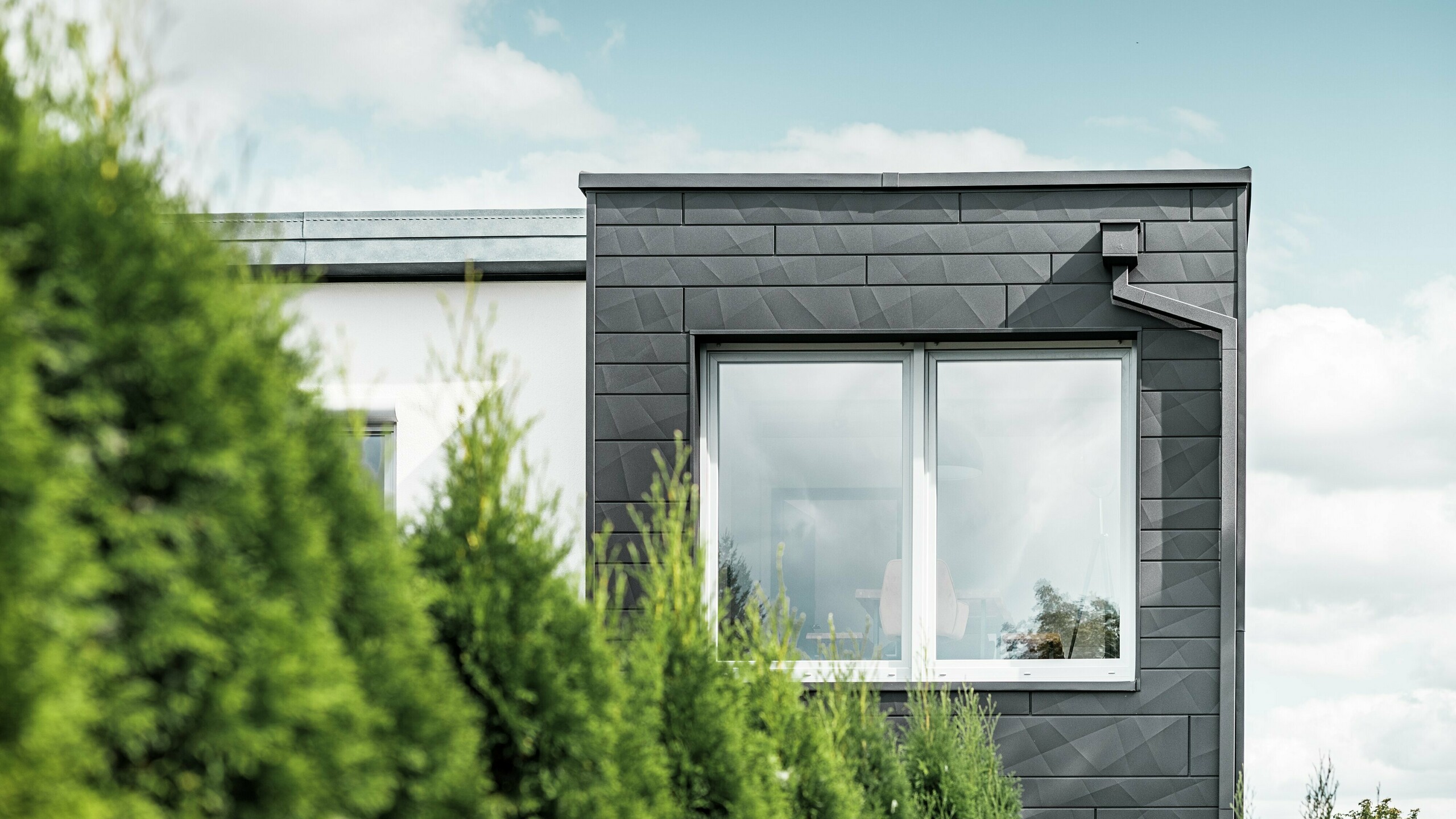 The clearly structured PREFA siding.X façade panels in colour P.10 black adorn the exterior wall of a single-family home in Teuschnitz, giving it a unique appearance. The PREFA roof drainage system, consisting of the square downpipe and the leader head, blends seamlessly into the design. The green thuja trees in the foreground contrast with the dark sheet metal facade.