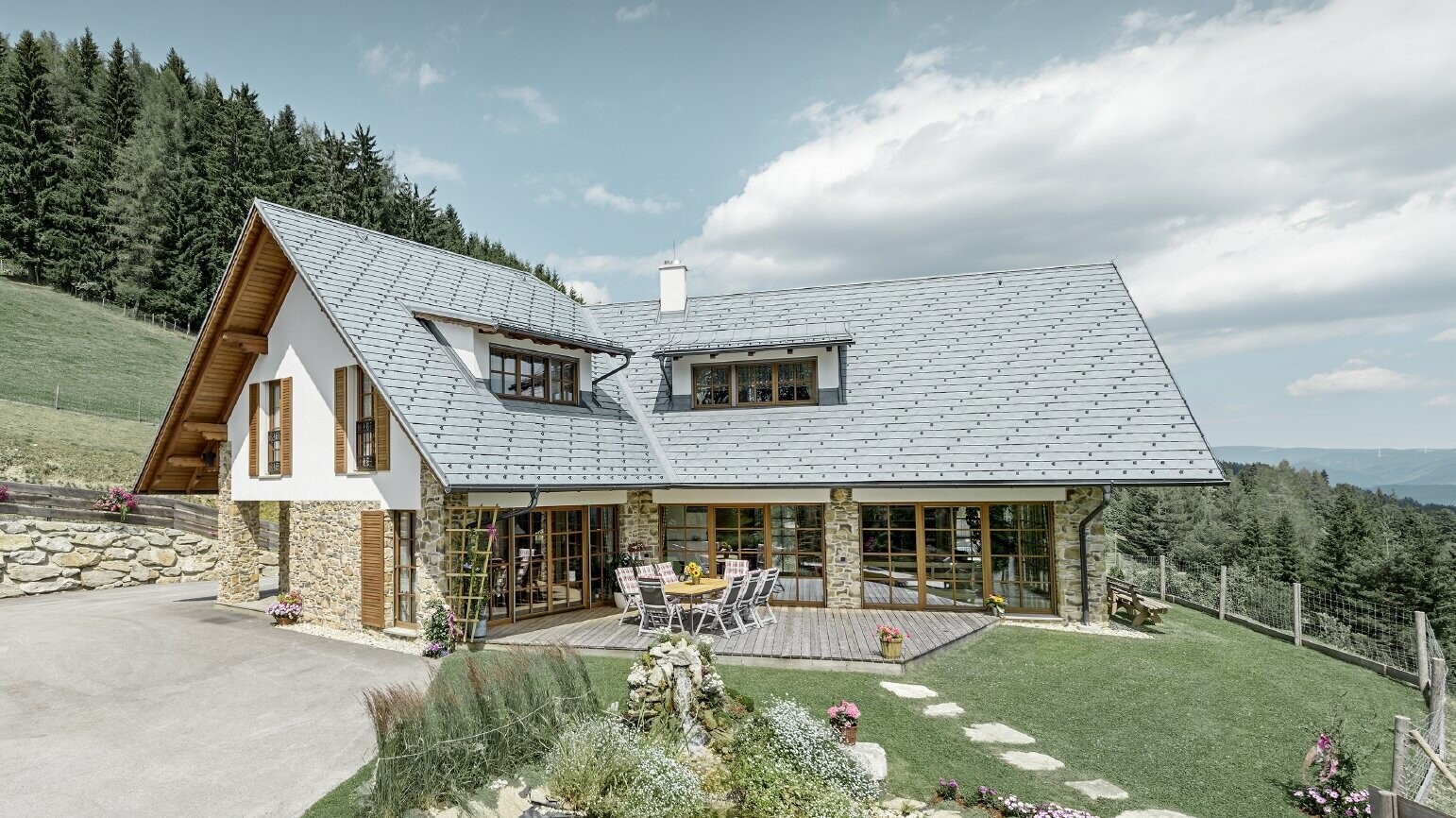 The detached house located on a slope, designed in an L shape, has a gabled roof with two eyebrow dormers. The roof was clad in PREFA roof shingles in stone grey. A lake is shown in the foreground and a forest can been seen in the background.