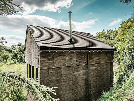 Blick von der Rückseite eines Hauses mit PREFA-Dachschindeln in der Farbe P.10 Walnussbraun und einem PREFA-Entwässerungssystem, umgeben von natürlichem Gelände im Westen Frankreichs.