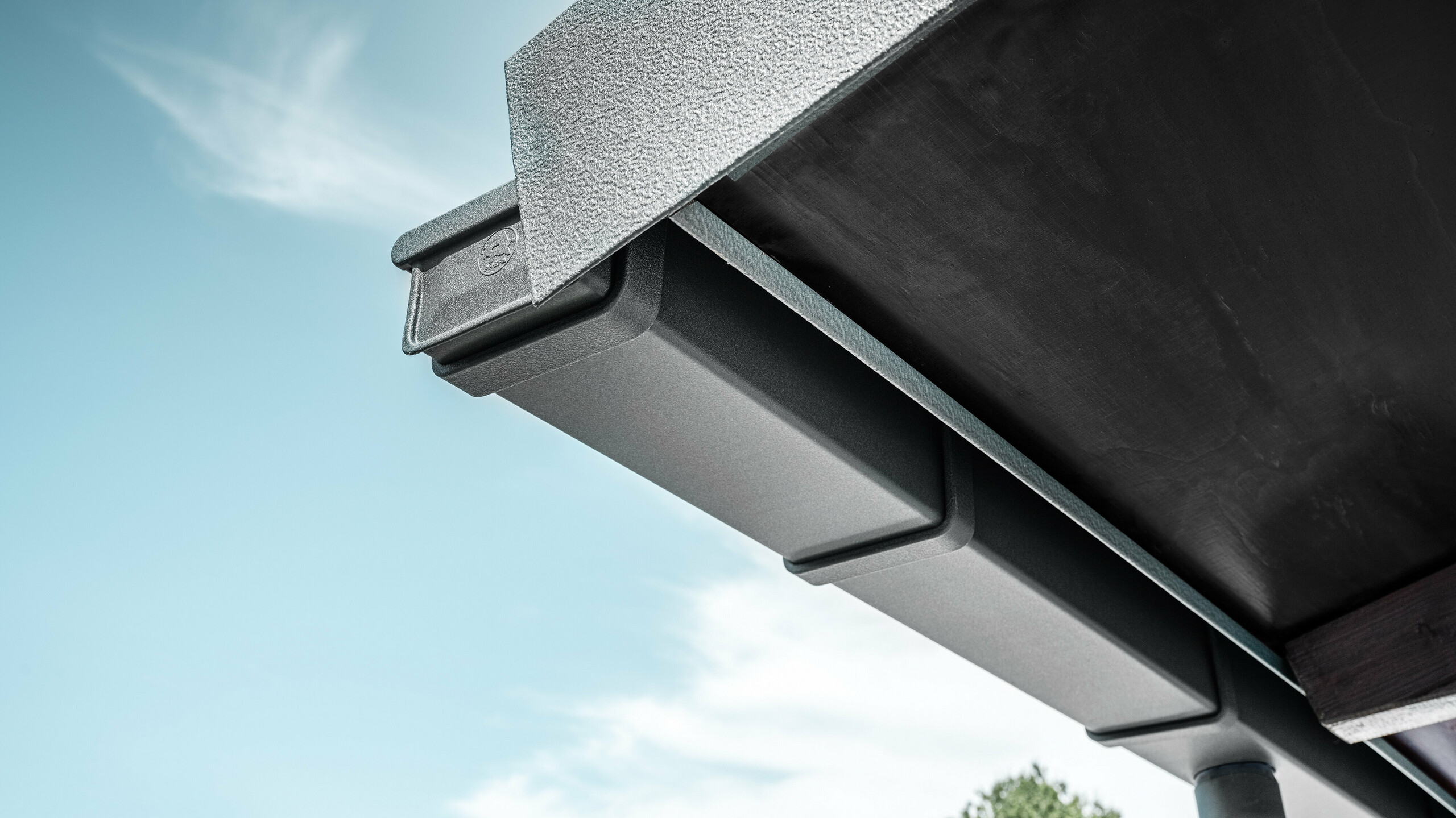 Detailaufnahme einer eckigen PREFA Kastenrinne in Anthrazit aus Aluminium mit strukturiertem Abschlussblech, montiert an der Dachkante eines Gebäudes vor blauem Himmel mit leichter Bewölkung.
