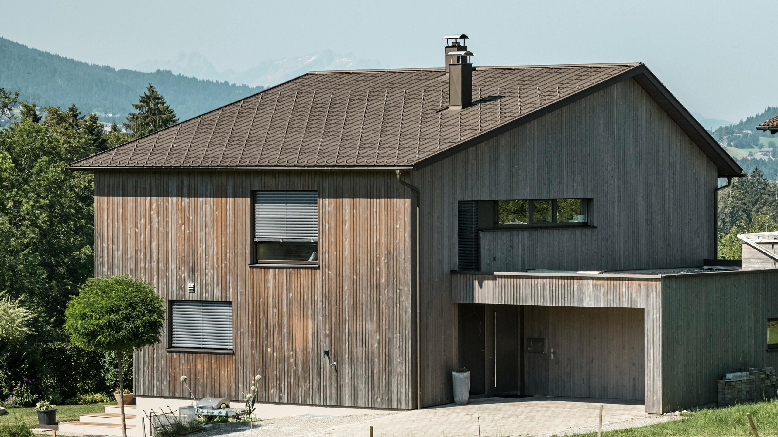 Side view of a contemporary detached house in Krumbach with a combination of a natural wooden façade and a long-lasting PREFA aluminium roof in P.10 brown. The harmonious colour scheme shows the elegant integration of modern design and traditional materials, with the PREFA roof tiles providing an aesthetic and functional roof design.