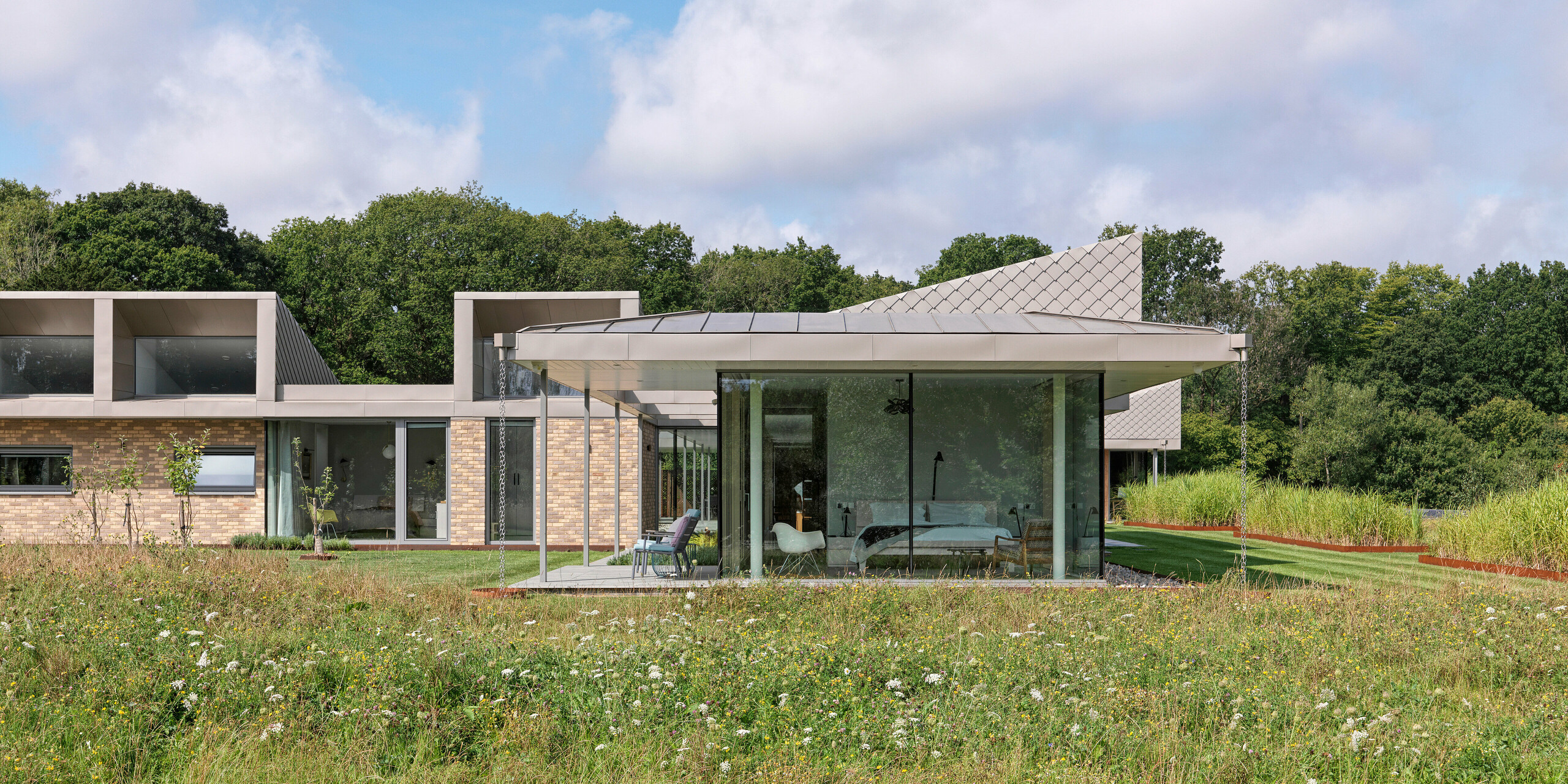 Side view of the modern bungalow in West Sussex with standing seam roofing and accentuated façade areas made of PREFA rhomboid façade tiles 20 × 20 in P.10 bronze.The floor-to-ceiling glass fronts of the living areas open onto a wildflower meadow in the foreground, creating a seamless connection between the interior and nature. The durable PREFA roof and façade system not only protects the building, but also lends it architectural elegance.