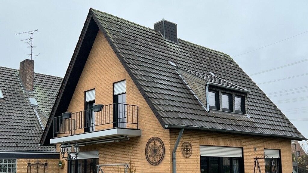 Detached house made of yellow clinker brick with a steep gable roof and dark roofing before the roof renovation; on the garden side with balcony, dormer window, terrace with seating and lawn in the foreground.