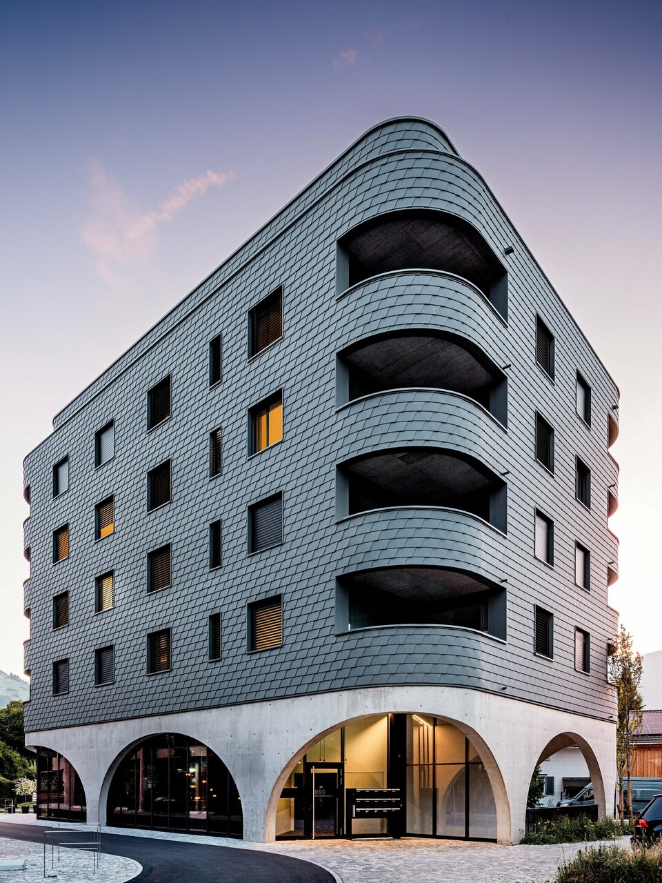 Modernes Mixed-Use-Gebäude in Buchs mit markant gerundeter, dreieckiger Form und durchgehender Fassade aus PREFA Wandschindeln in P.10 Hellgrau. Die schuppenartige Aluminiumhaut umhüllt das Gebäude wie eine Reptilienstruktur und betont die fließenden Rundungen sowie die tief eingeschnittenen Balkone. Über dem massiven Sockel mit eleganten Betonarkaden wirkt die leichte, witterungsbeständige PREFA Fassade besonders ausdrucksstark. Präzise verlegte Aluminiumschindeln sorgen für eine homogene, langlebige und wartungsarme Gebäudehülle mit starkem Wiedererkennungswert.