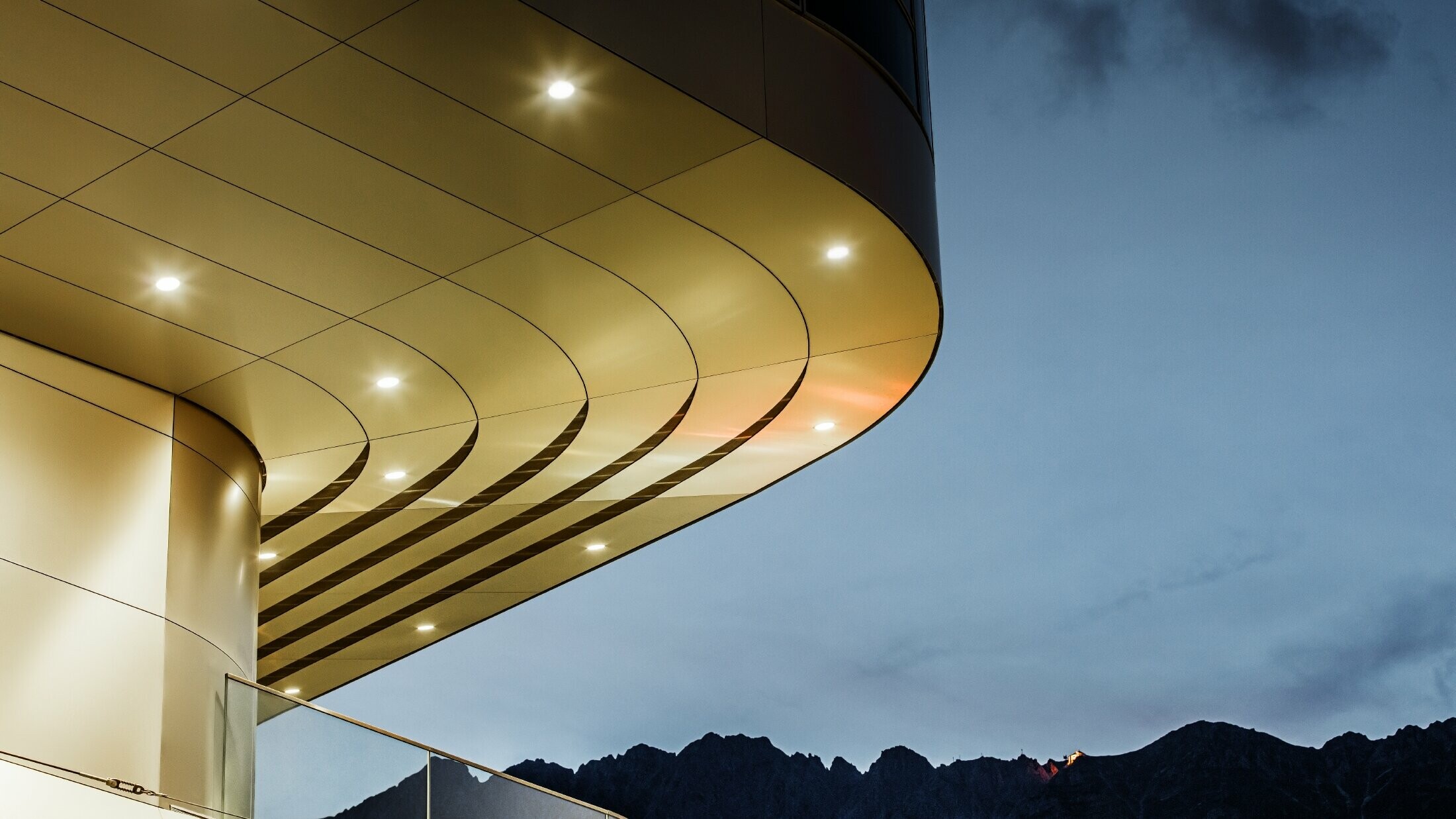 The image shows an impressive architectural detail view of a PREFA PEMA composite panel made of brushed aluminium. The panel is gently curved and flows in elegant, flowing lines, which are particularly accentuated by targeted lighting in the evening. The clear structure and the elegant aluminium look of the panel complement the modern design of the façade and offer an elegant, contemporary solution for high-quality architecture.