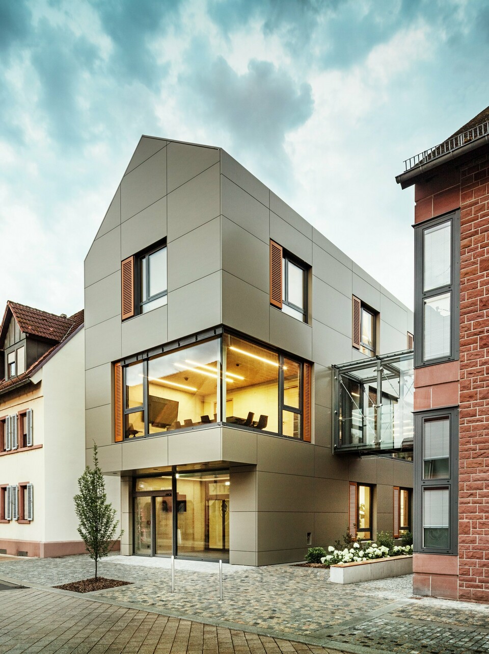 The image shows the modern town hall in Alzenau, which is clad in aluminium composite panels from PREFA in a warm bronze tone. The façade is interrupted by large windows that offer a view of the interior, which is illuminated by warm light. The glass walkway on the right-hand side of the picture connects the building to a neighbouring traditional brick building, creating an exciting contrast between modern and historic architecture.