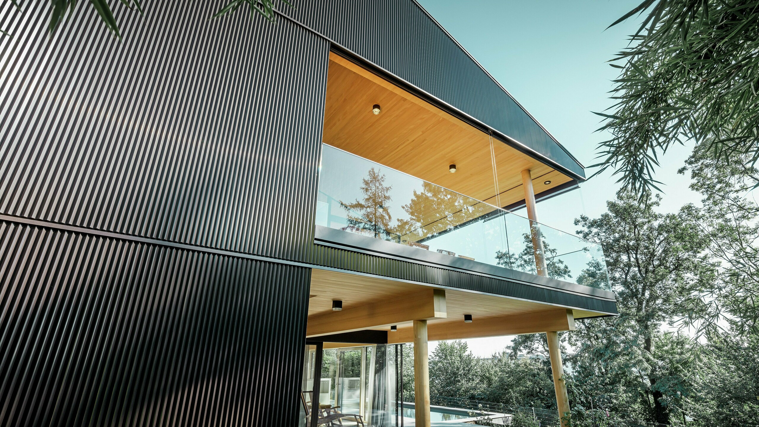 Side view of a detached house, the façade is clad with dark PREFA serrated profile. You can see the terrace and a spacious balcony. The ceiling or soffit is designed with wooden panels.