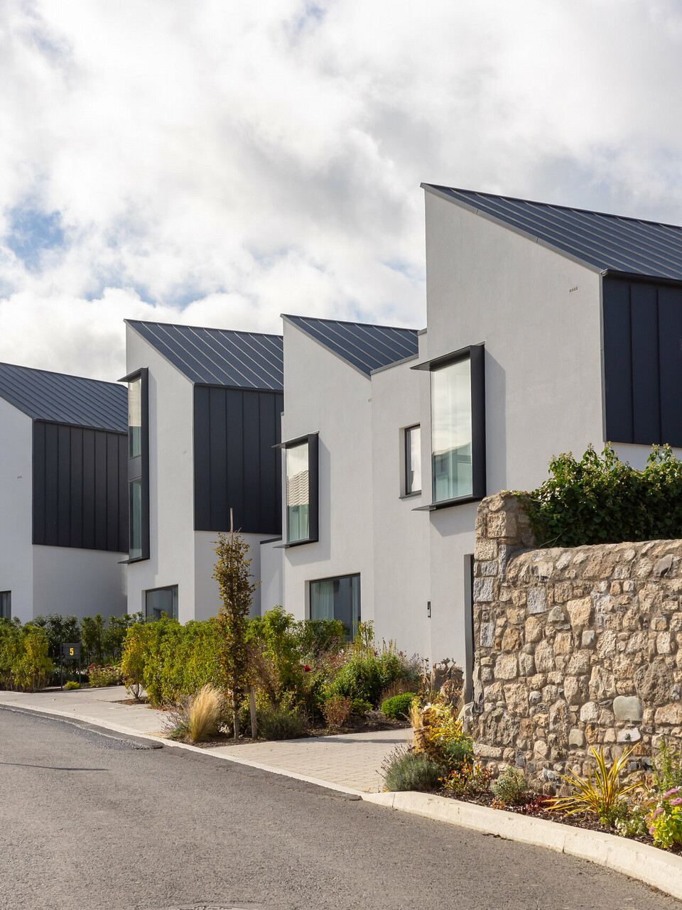 Watson Place terraced housing development with striking sloping roofs and continuous PREFALZ standing seam cladding in P.10 anthracite. The dark aluminium surfaces create a striking contrast to the light-coloured façades and natural stone walls. The durable PREFA aluminium provides reliable weather protection and gives the modern architecture a clear, high-quality appearance.
