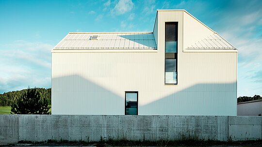 Das moderne Einfamilienhaus in Freistadt wurde aus einer seitlichen Perspektive fotografiert, um das markante Design der Fassade und des Daches hervorzuheben. Die reinweiße PREFALZ-Aluminiumverkleidung in Kombination mit dem steilen Satteldach und den schmalen Fenstern verleiht dem Gebäude eine moderne und zeitlose Ausstrahlung. Die vertikalen Rillen der Fassade verleihen dem Gebäude eine dynamische, texturierte Erscheinung. Das Lichtspiel, das durch die Sonne auf die Fassade fällt, sorgt für interessante Schatteneffekte und betont die präzisen Linien des Designs. Diese klare und minimalistische Architektur wird durch die langlebige und witterungsbeständige PREFALZ-Fassade unterstrichen, die für ihre Haltbarkeit und Pflegeleichtigkeit bekannt ist.