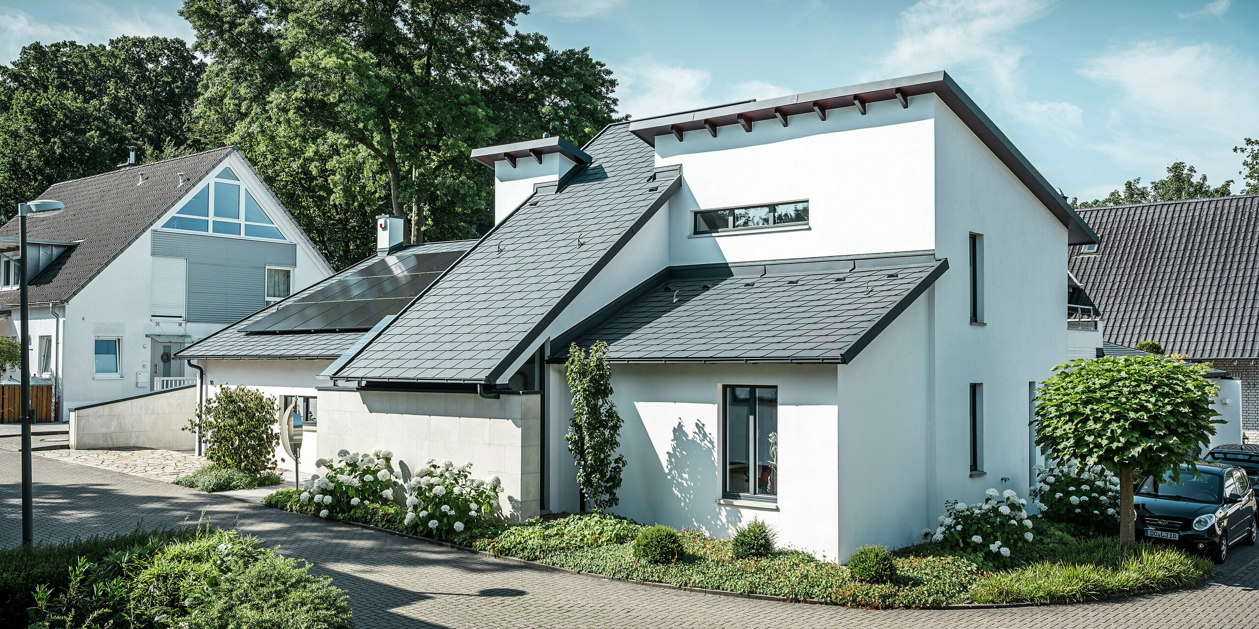 Modern detached house with white plaster façade and dark PREFA aluminium pitched roof. Integrated photovoltaic modules and anthracite aluminium roof drainage can be seen on the roof. The building is located in a well-maintained residential area with planted front gardens, paved driveways and surrounding detached houses.
