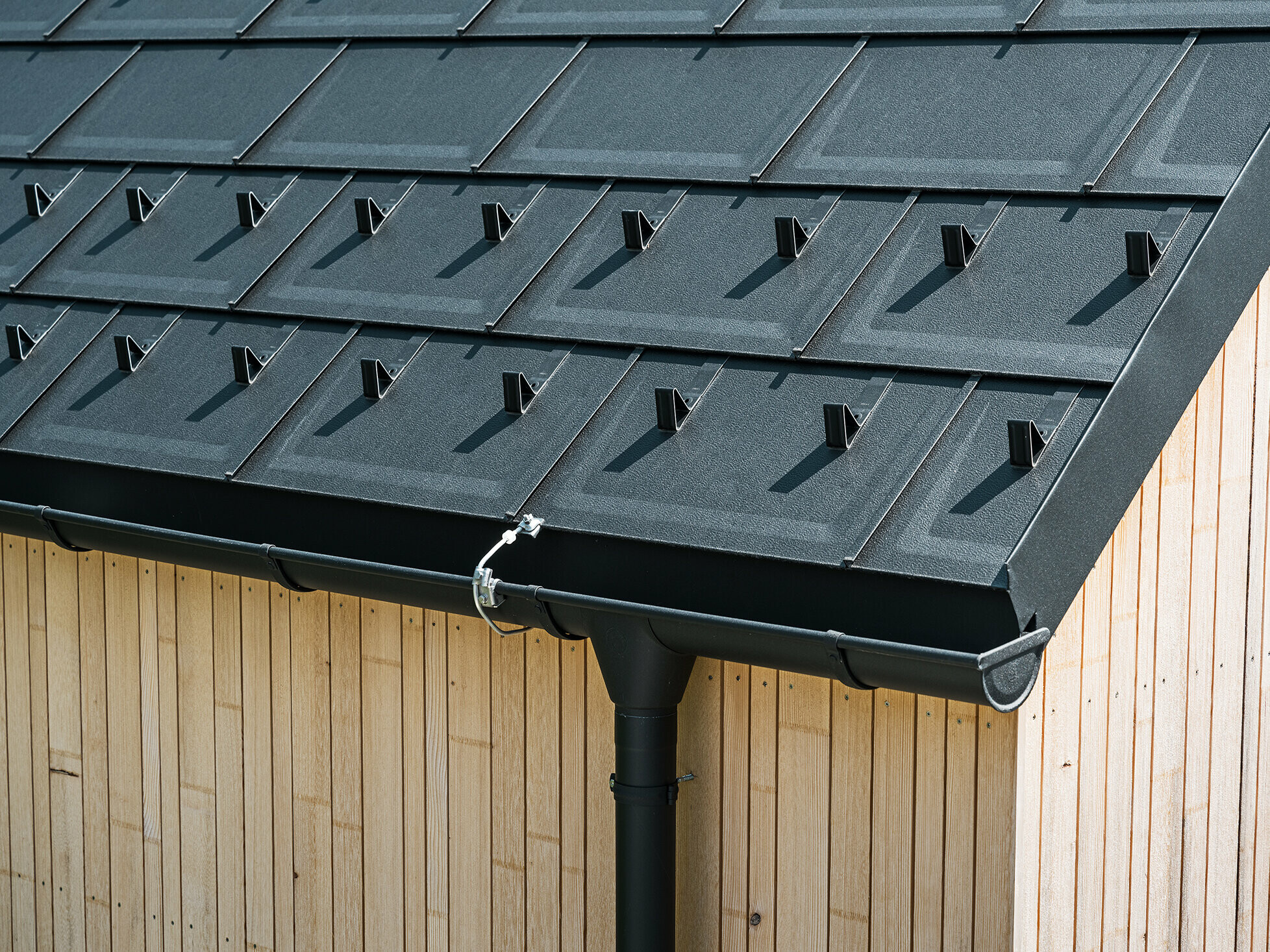 Close-up of PREFA roof tile R.16 with snow guards, gutter and downpipe in black P.10 incl. lightning protection and a vertical wooden façade