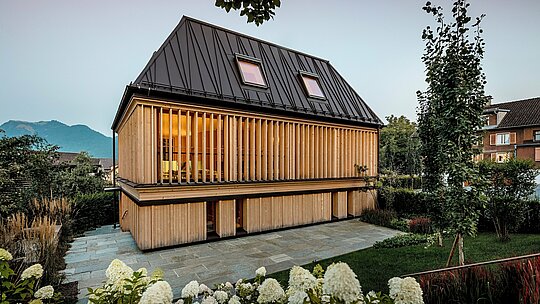 Das moderne Badehaus des Hotels Hirschen in Schwarzenberg verfügt über ein preisgekröntes PREFA Stehfalzdach in der Farbe P.10 Nussbraun. Das ikonische Walmdach mit 60-Grad-Neigung und unkonventionell gesetzten Stehfalzen ist ein architektonisches Highlight, das Tradition und Innovation verbindet. Die Fassade aus vertikalen Holzlamellen verleiht dem Gebäude eine warme, natürliche Ausstrahlung und fügt sich harmonisch in den denkmalgeschützten Ortskern ein. PREFALZ überzeugt durch Langlebigkeit, Formfreiheit und exzellente Verarbeitbarkeit, auch bei komplexen Dachformen. Ein preisgekröntes Beispiel für innovative Architektur mit hochwertigen PREFA-Produkten.