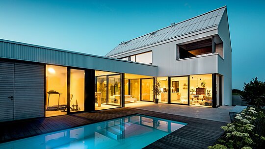 Das Bild zeigt das moderne Einfamilienhaus in Freistadt mit seiner markanten PREFALZ-Fassade in Reinweiß. Die präzise gefaltete Aluminiumfassade verleiht dem Gebäude eine klare, geometrische Eleganz, die sich harmonisch mit den großzügigen Fensterflächen und dem Flachdach kombiniert. Die Verwendung von PREFALZ sorgt nicht nur für ästhetische Beständigkeit, sondern auch für eine hohe Witterungsbeständigkeit und Langlebigkeit. Der offene Innenbereich mit Blick auf den Pool wird durch die klaren Linien und das minimalistische Design perfekt ergänzt, wodurch das Haus eine zeitgemäße, nachhaltige Wohnlösung darstellt.