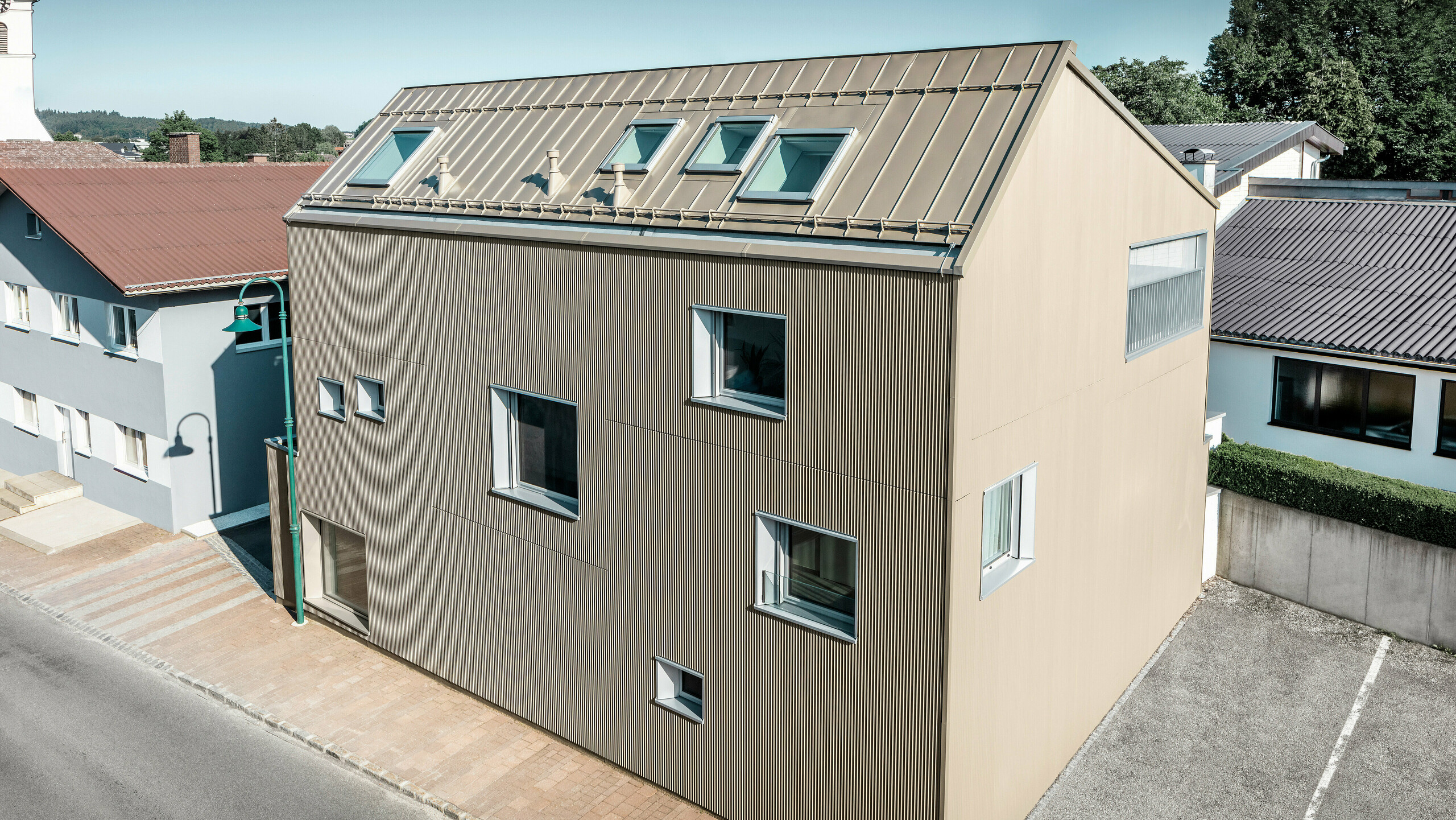 Bird's-eye view of a detached house in Frankenburg, Austria, which stands out thanks to its PREFA façade in the form of a serrated profile and a PREFALZ roof, both in a matching bronze colour. The façade has a striking texture that captures natural light and creates a lively play of shadows. Several roof windows allow natural light to flood into the internal space and emphasise the functional and aesthetic integration of modern architectural elements. With its contemporary design, the house blends harmoniously into the typical Austrian village environment while at the same time providing a contrasting accent to the surrounding traditional buildings.