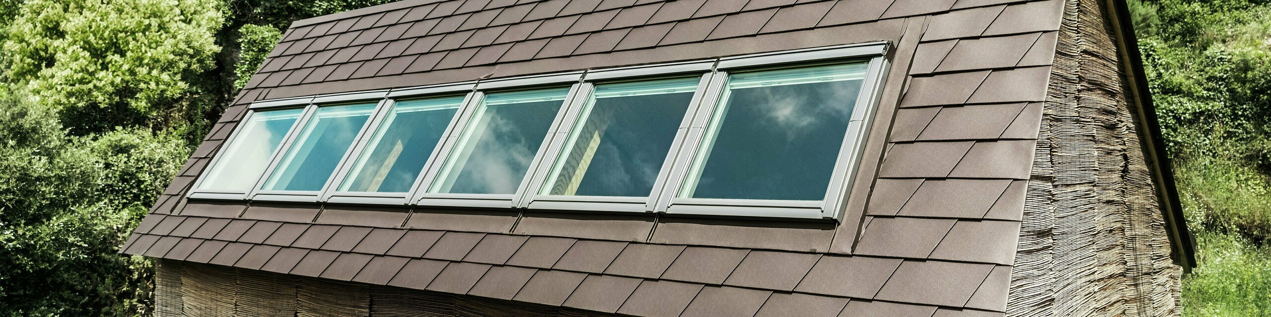 PREFA Dachschindeln in der Farbe P.10 Walnussbraun auf dem Dach eines modernen Hauses mit Dachfenstern – eine langlebige Aluminiumlösung für Energieeffizienz und stilvolles Dachdesign.